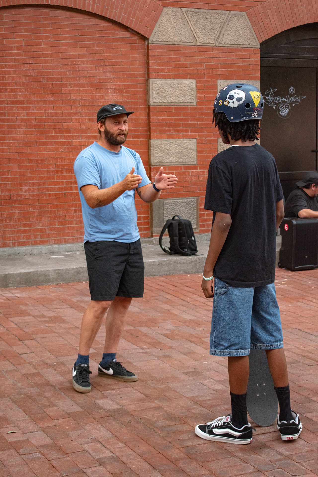 A STOKED coach in a blue tee talking with his hands to a young person in a helmet, both standing on brick paving outside a skate spot.