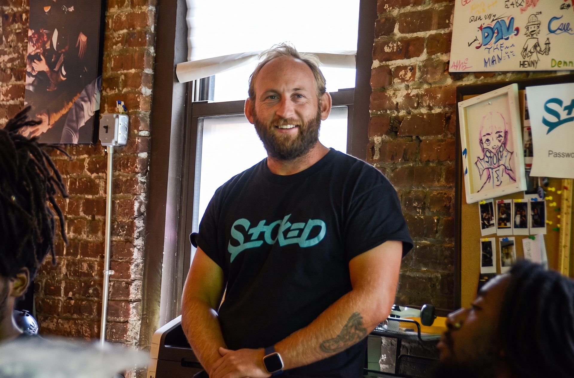 A veteran STOKED coach in a black tee, smiling mid conversation with two younger coaches in the brick-walled STOKED office.