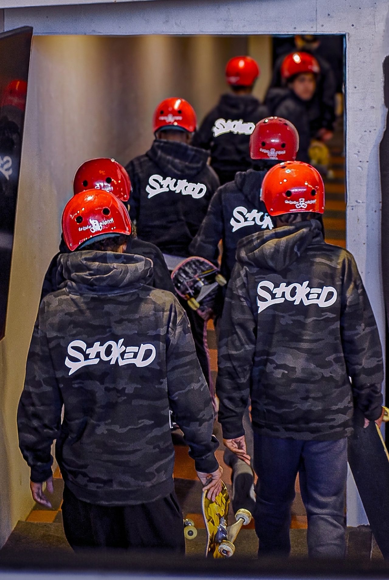 A crew of young people in matching red helmets and camo STOKED hoodies, boards in hand, heading in to a session together, seen from behind.