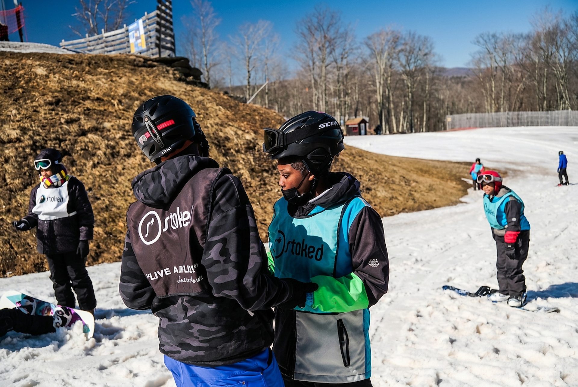 A STOKED coach helping a young snowboarder in a teal STOKED bib with their gear, one to one, at the base of a beginner slope.
